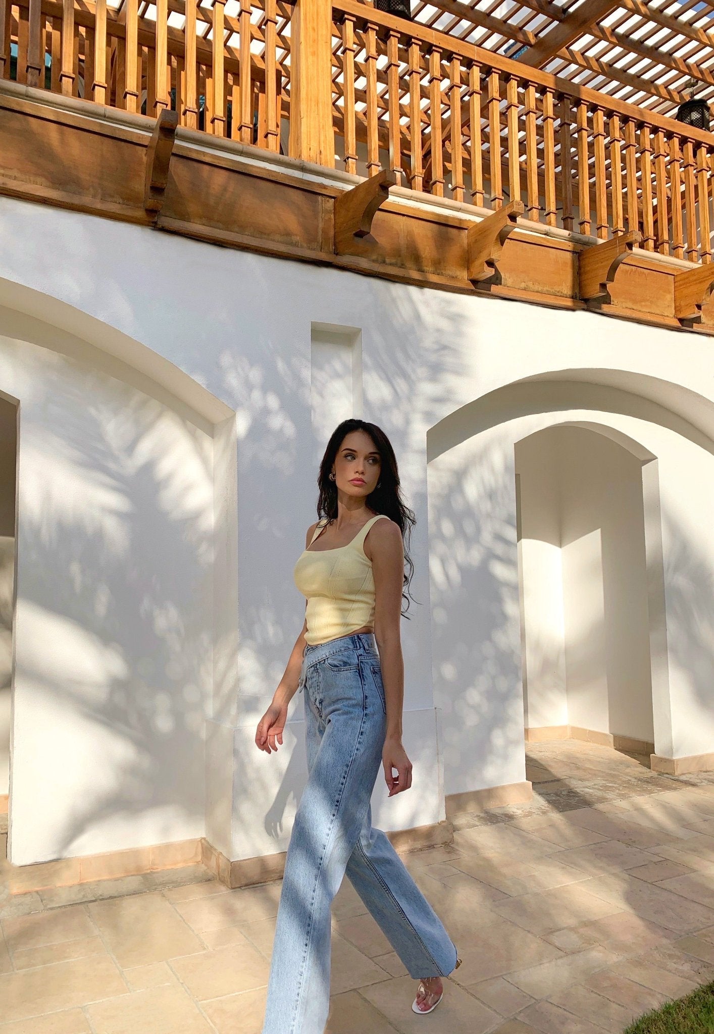 Wearing the Anastacia Jeans with an asymmetric belt, a woman strides confidently under a wooden pergola in a yellow top. Leaf shadows are artistically cast on the white wall behind her, perfectly complementing her vintage-inspired style and the serene, sunlit ambiance.