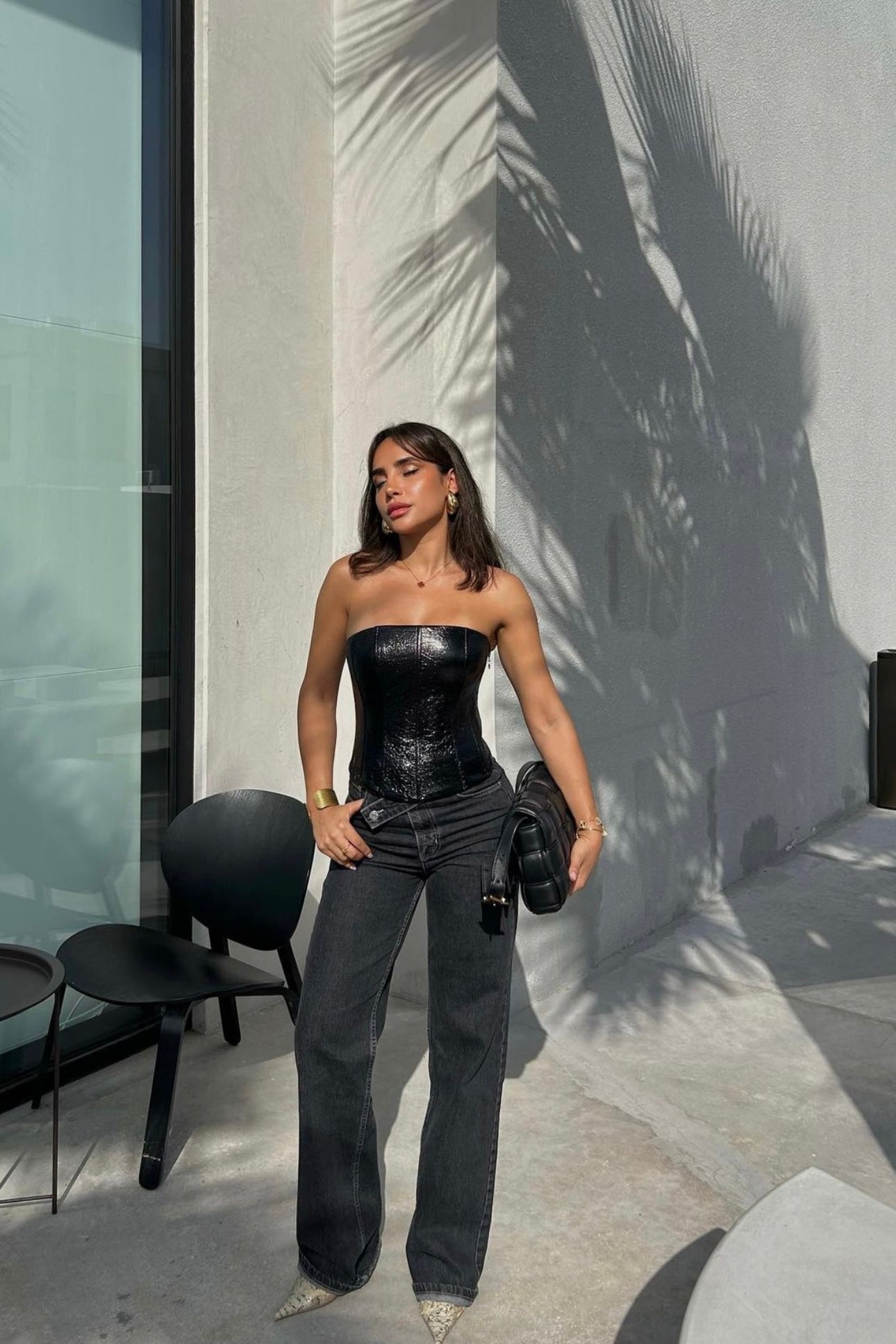 A woman stands confidently outside a building with palm shadows on the wall. She wears a black corset top paired with Anastacia Jeans featuring an asymmetric belt, and she holds a black handbag. Nearby, a black chair and vintage-inspired table accentuate the scene, with sunlight casting long shadows.