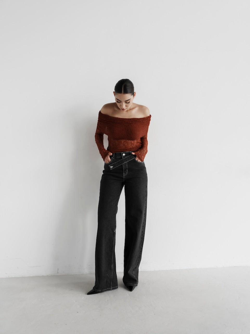 A person with dark hair stands against a white wall, looking down. They wear a rust-colored off-shoulder top paired with the Anastacia Jeans featuring an asymmetric belt, creating a vintage-inspired look. The setting is minimalist and well-lit.