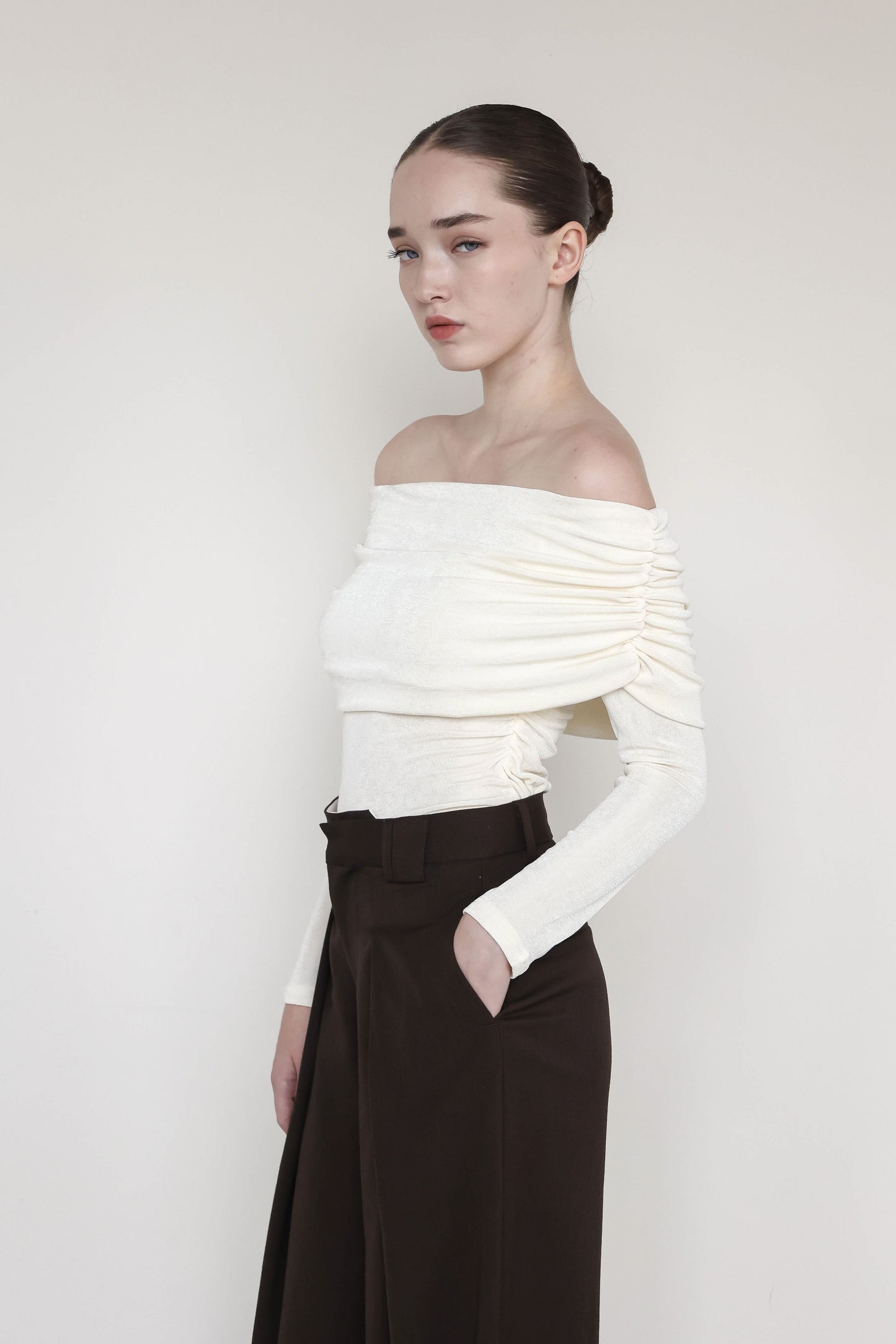 A young woman is wearing an elegant off-the-shoulder cream-colored top with ruched detailing along the sleeves. Her hair is styled in a sleek, braided bun, and she stands slightly turned, with her head bowed and hand in her pocket. She is paired with high-waisted, wide-leg brown trousers, creating a sophisticated and minimalist look against a neutral background. The soft, flowing lines of the outfit highlight a graceful and contemplative pose.