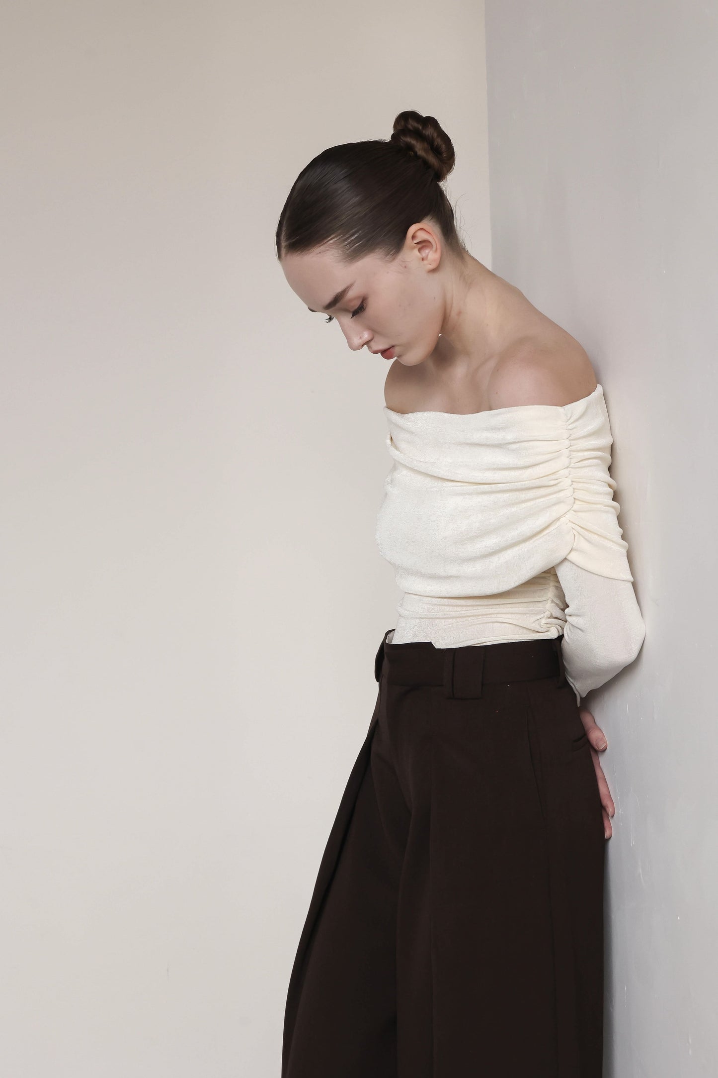 A young woman is wearing an elegant off-the-shoulder cream-colored top with ruched detailing along the sleeves. Her hair is styled in a sleek, braided bun, and she stands slightly turned, with her head bowed and hands behind her back. She is paired with high-waisted, wide-leg brown trousers, creating a sophisticated and minimalist look against a neutral background. The soft, flowing lines of the outfit highlight a graceful and contemplative pose.