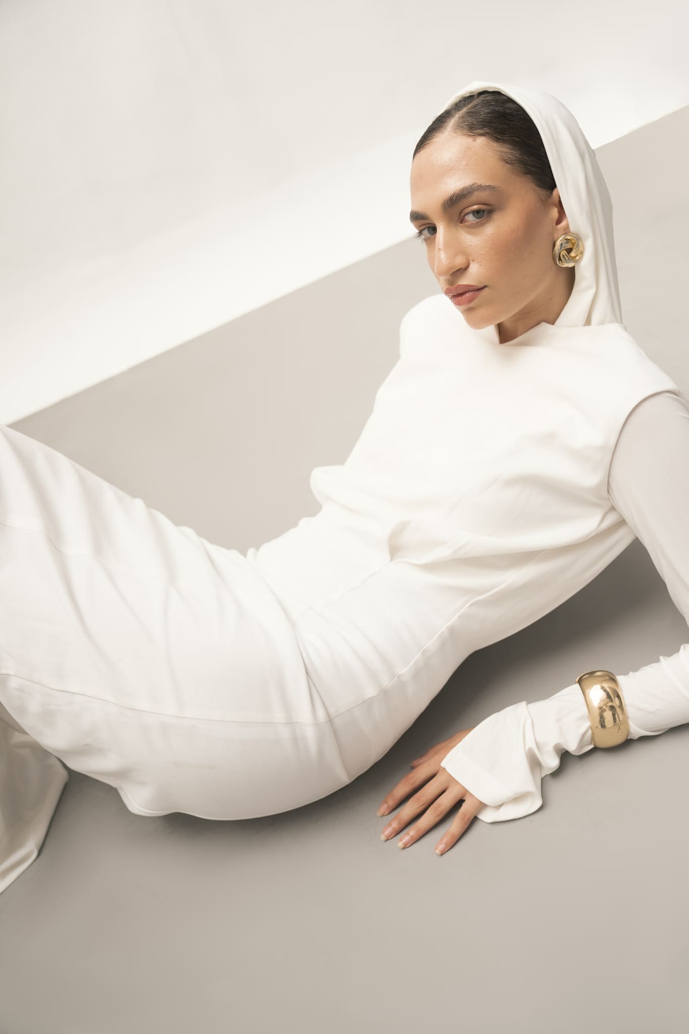 A woman poses gracefully on a light gray floor, highlighting the minimalist elegance of the Avelia Dress in fitted white jersey knit. Her look is enhanced by long sleeves and a hood, with large gold earrings and a chunky gold bracelet adding opulence. Her gaze remains calm and focused.