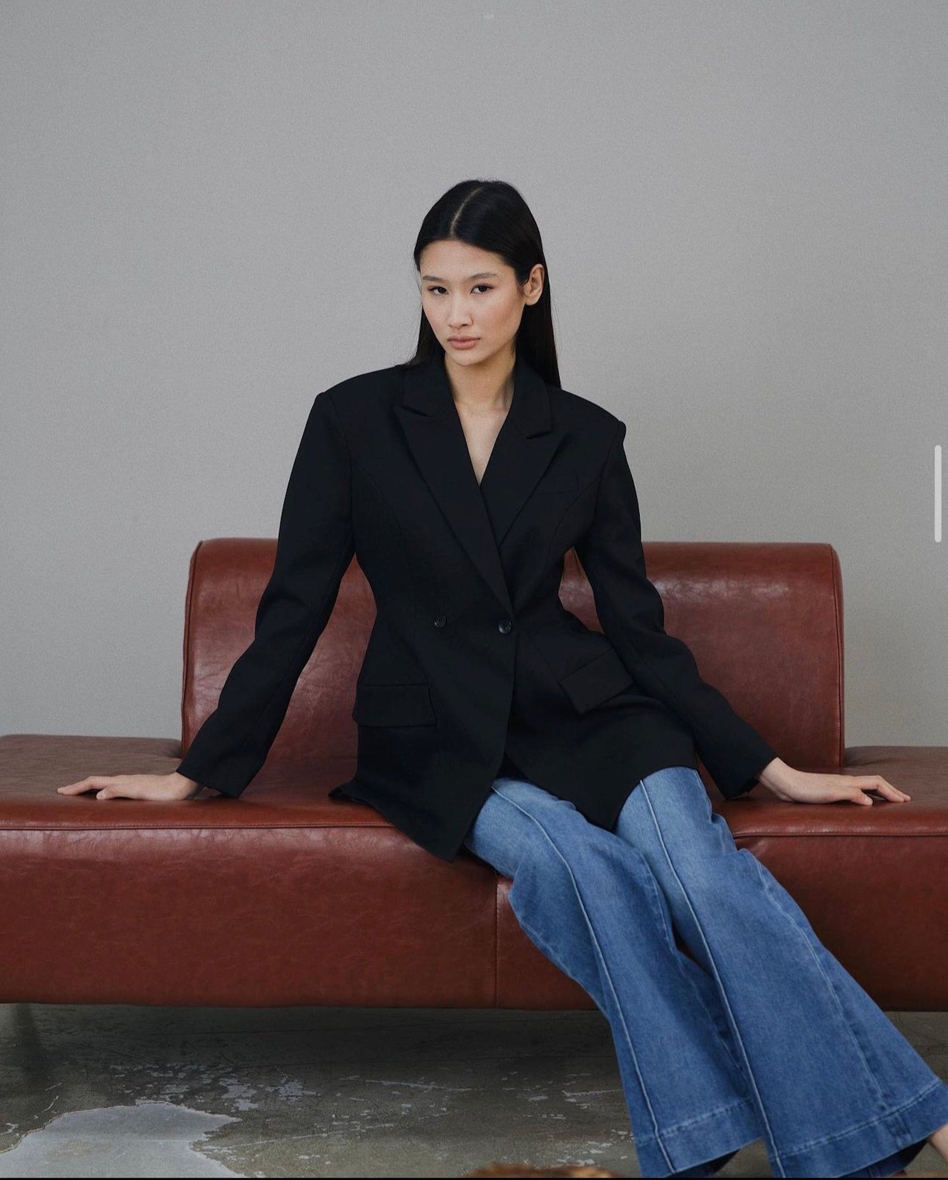 A person with long dark hair sits on a brown leather bench, wearing the Olivia tailored blazer with a double-breasted design and blue flared jeans. They maintain a neutral expression against a plain gray wall, seated on a concrete floor.